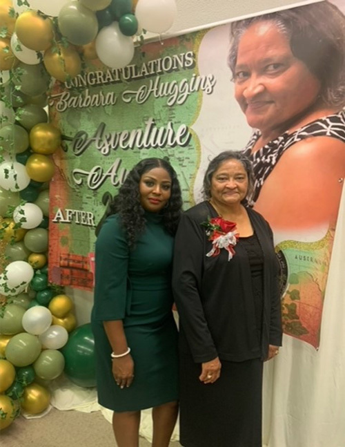 Barbara Huggins and another woman stand together in front of a banner that says: Congratulations Barbara Huggins. Adventure Awaits.